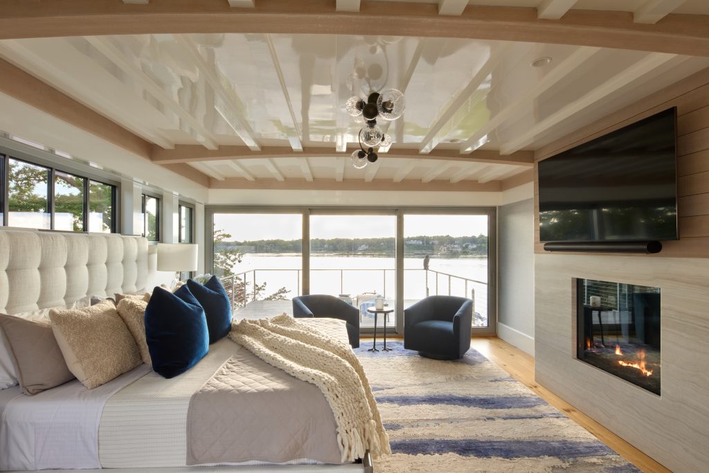 Coveledge bedroom with a bed, two sofa chairs, a balcony, a tv mounted on the wall with a fireplace underneath